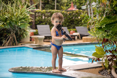 Full length of girl holding camera standing by swimming pool