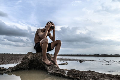 Sad boy sitting on rock by sea against cloudy sky