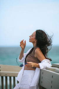 Side view of young woman using mobile phone while sitting at beach against clear sky