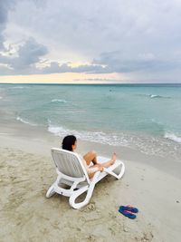 High angle view of woman looking at view while resting on lounge chair at beach against cloudy sky