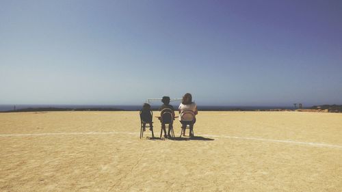 Rear view of mother sitting with children on chairs against sky at sandy beach during sunny day