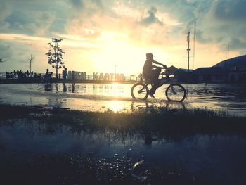 Silhouette man by bicycle on lake against sky during sunset