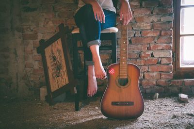 Low section of woman with acoustic guitar sitting on chair against brick wall