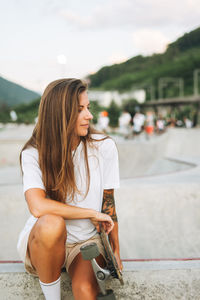 Slim young woman with long blonde hair in light sports clothes with longboard in outdoor skatepark 