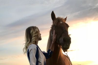 Low angle view of woman petting horse against cloudy sky during sunset