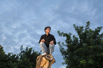 Young transgender man with glasses posing on a log outdoors.