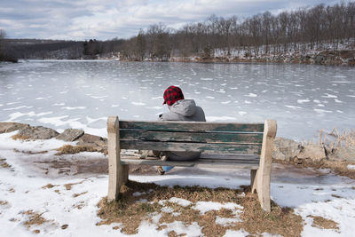 Man sitting on bench by frozen lake against sky during winter