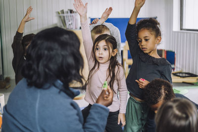 Female teacher asking questions and students raising hand to answer at day care center