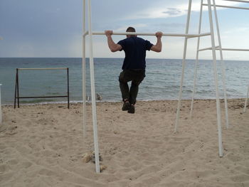 Rear view of mid adult man hanging from metallic structure at sandy beach
