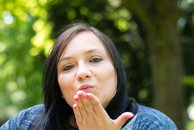 Close-up portrait of woman blowing kiss against trees at park