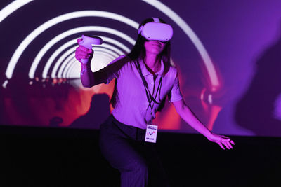 Young female entrepreneur holding joystick watching through vr glasses at illuminated workshop