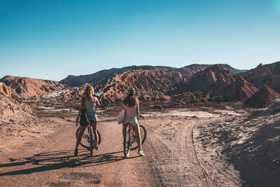 Rear view of friends riding bicycles on field against clear blue sky during sunny day