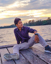 Young man sitting on wood against lake during sunset