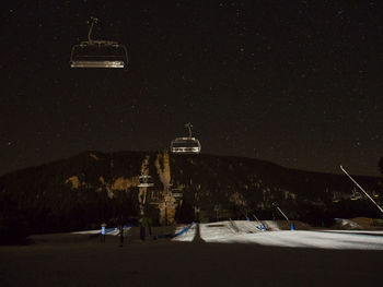 Ski lifts over snow covered field by mountain at night
