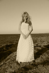 Sad young woman wearing white dress standing at beach against sky