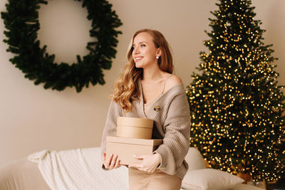 Portrait of a smiling woman in a cardigan holding christmas gift boxes in a decorated house