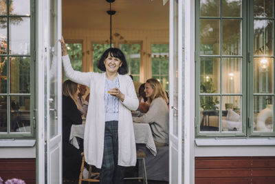 Portrait of woman wearing cardigan holding drink glass while standing at doorway