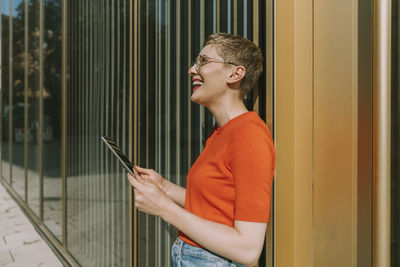 Woman laughing while holding digital tablet standing against building exterior on sunny day