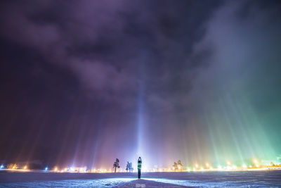 Rear view of person with arms raised standing at illuminated beach against sky at night