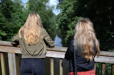 Rear view of sisters standing by railing on footbridge over river against trees