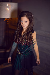 Girl in a green figure skating dress stands next to a chair and a lamp in the room