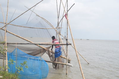 Man working on fishing net in sea against sky