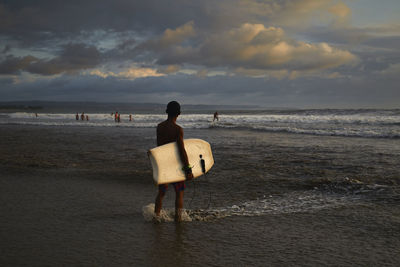 Rear view of man standing on shore while holding surfboard during sunset