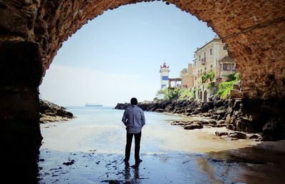 Rear view of man standing below arch at beach against sky