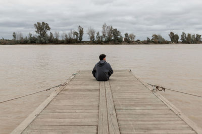 Rear view of man sitting on pier over lake against sky