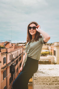 A young smiling woman in sunglasses sits on a rooftop with a panoramic view of rome, italy