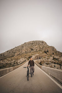 Young man wheeling bicycle on road against mountain