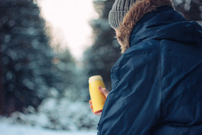 Side view of man having drink while sitting against trees during winter