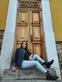 Full length portrait of smiling young woman sitting against door