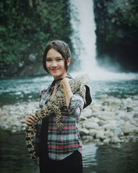 Portrait of smiling young woman holding crocodile against waterfall