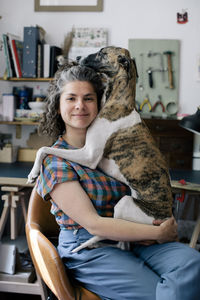 Portrait of smiling woman embracing dog while sitting on chair at home