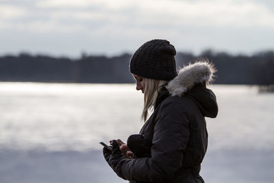 Side view of woman in hooded shirt and knit hat using phone standing by lake