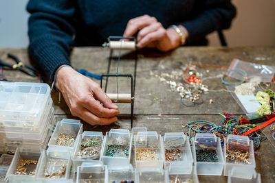 Crop anonymous person using professional instrument with leather cord while making bijouterie at table with various decorative elements in workshop