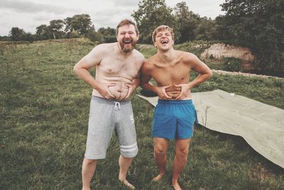 Shirtless cheerful father and son holding abdomen on grassy field