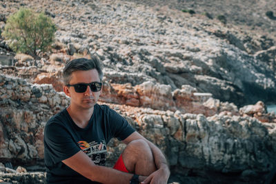 A man sits and hugs his knee against the backdrop of the rocky mountains.