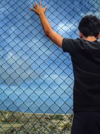 Boy standing by chainlink fence against clear sky