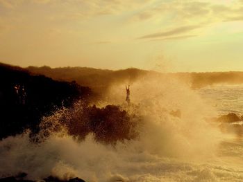 Waves splashing at rocky shore against cloudy sky during sunset