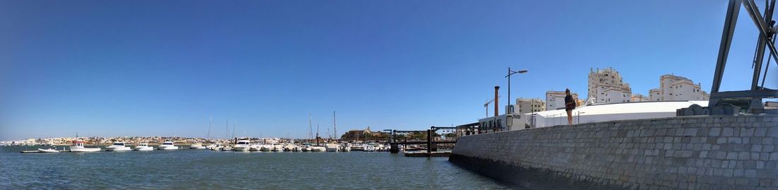 Panoramic shot of woman standing at harbor by sea against clear blue sky