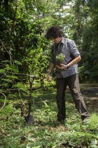 Hispanic man planting a small plant with a black shovel in a green field surrounded by trees