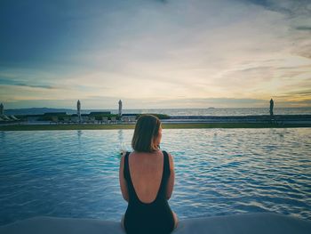 Rear view of woman looking away while sitting by swimming pool at sunset