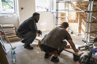 Multiracial male carpenters crouching in front of plank while in apartment