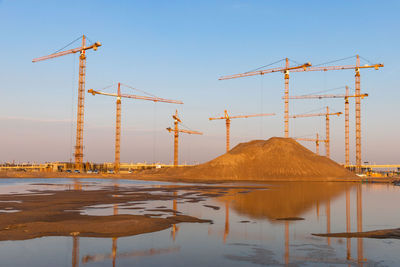 High pile of sand and group high-altitude tower cranes at construction site, sunset sky background.