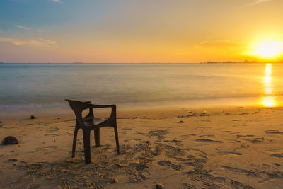 Empty chair at beach against sky during sunset