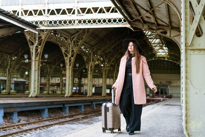 Beautiful young casual tourist woman with a suitcase in a pink coat waiting