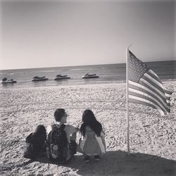 Rear view of people by american flag sitting at beach against clear sky