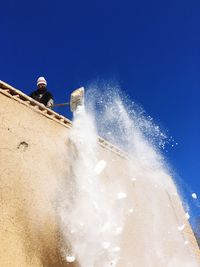 Low angle view of person throwing snow from roof against blue sky